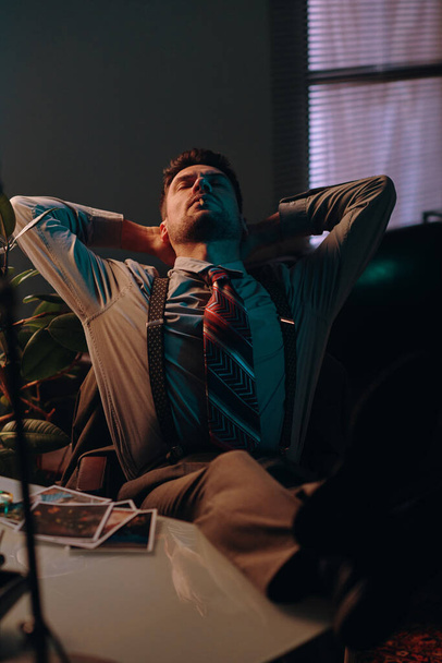 Νεαρός άνδρας με ρετρό ρούχα καπνίζει ενώ χαλαρώνει στο χώρο εργασίας στο σκοτεινό γραφείο στο διάλειμμα με τα χέρια του πίσω από το κεφάλι - Φωτογραφία, εικόνα