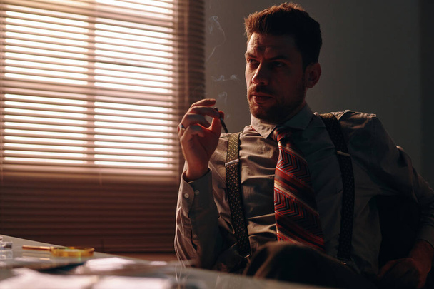 Молодой задумчивый человек в ретро-формальной одежде думает о чем-то и курит сигарету, сидя за столом в темном кабинете - Фото, изображение