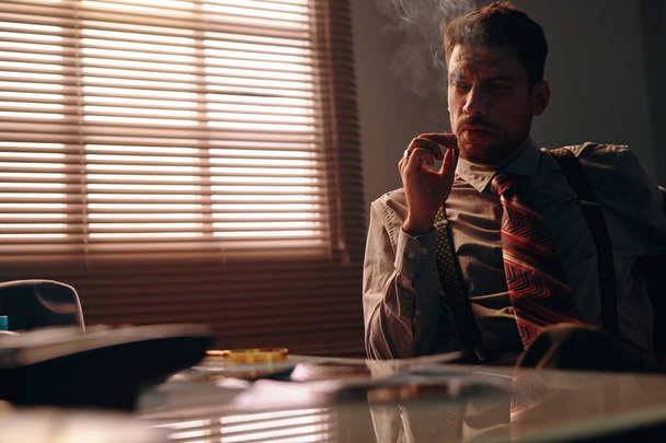 Fiatal, komoly üzletember formalwear dohányzik cigaretta, miközben ül a munkahelyén sötét irodában ablakkal borított velencei vak - Fotó, kép
