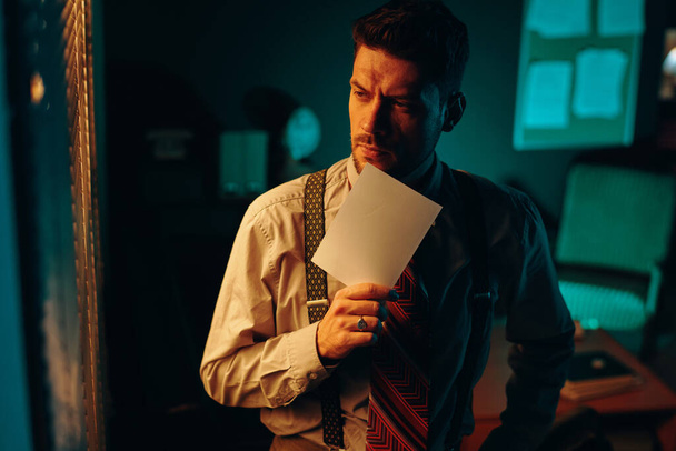 Jeune officier ou détective masculin rétro coûteux avec photo de son visage debout devant la caméra dans un bureau sombre et regardant par la fenêtre - Photo, image