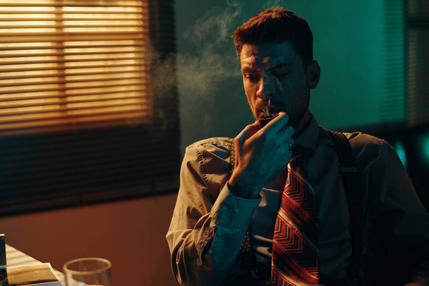 Töprengő fiatalember dohány pipa a szájban ül a munkahelyen a kamera előtt munka közben sötét irodában vakokkal az ablakon - Fotó, kép