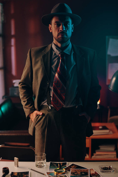 Junger Mann in altmodischem Anzug und Hut steht vor der Kamera an seinem Arbeitsplatz mit Bildern im dunklen Büro und schaut einen an - Foto, Bild