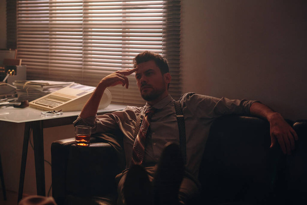 Joven oficinista pensativo con vaso de whisky relajante en cómodo sofá de cuero negro después del trabajo y pensando en algo - Foto, Imagen