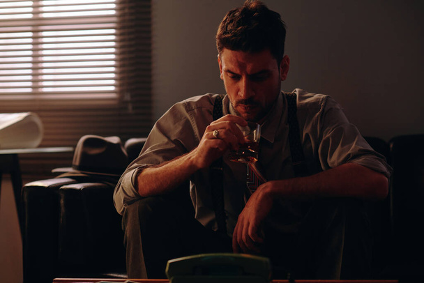 Серьезный бизнесмен смотрит на виски в стакане, сидя на диване в темном кабинете с окном, покрытым жалюзи - Фото, изображение