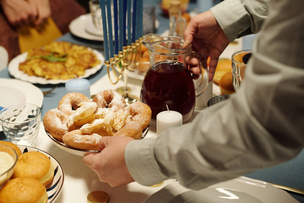 Ręce Żyda z dzbankiem soku żurawinowego i pączkami domowej roboty kładąc je na stole serwowane z apetycznym jedzeniem na obiad - Zdjęcie, obraz