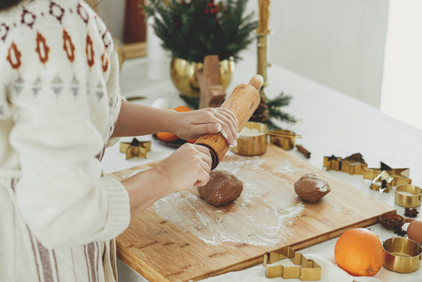 Mãos amassando massa de gengibre em tábua de madeira com rolo de pino, cortadores de metal dourado, especiarias culinárias, decorações festivas na cozinha branca moderna. Mulher fazendo biscoitos de gengibre de Natal - Foto, Imagem
