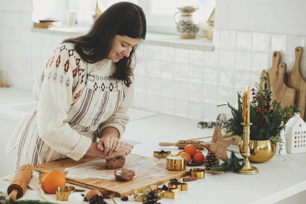 Женщина делает рождественское пряничное печенье на современной белой кухне. Ручное смешивание пряничного теста на деревянной доске с мукой, катком, золотистыми металлическими резаками, пряностями для приготовления пищи на столешнице - Фото, изображение