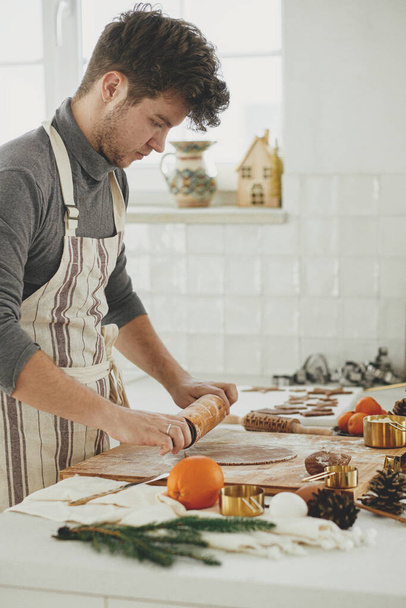 Man making christmas gingerbread cookies in modern white kitchen. Hand kneading gingerbread dough on wooden board with flour, rolling pin, golden metal cutters, cooking spices on countertop - Photo, Image