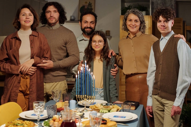 Sechsköpfige Familie verschiedener Generationen steht mit brennenden Menorah-Kerzen und hausgemachtem Essen am gedeckten Tisch und schaut einen an - Foto, Bild