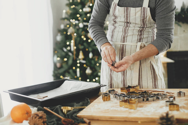 Ruce držící syrovou perníkovou sušenku ve slavnostních tvarech u tácu na rustikálním stole s vánoční výzdobou. Muž dělá vánoční perníkové sušenky, atmosférické svátky - Fotografie, Obrázek