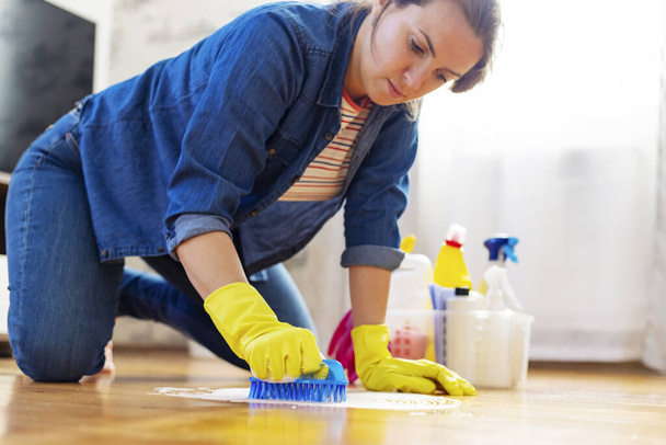 Μια νεαρή γυναίκα με προστατευτικά γάντια πλένει το πάτωμα με ένα πινέλο και απορρυπαντικό. Υπηρεσία καθαριότητας. Έννοια της οικιακής εργασίας και της οικιακής οικονομίας - Φωτογραφία, εικόνα