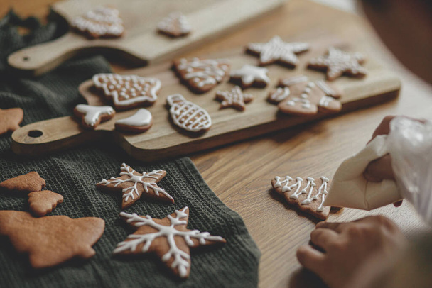 Eller, Noel kurabiyelerini kırsal ahşap masa üzerindeki kremayla süslüyor. Atmosferik Noel gelenekleri. Kurabiyeleri şekerli süslemek. Aile zamanı. - Fotoğraf, Görsel