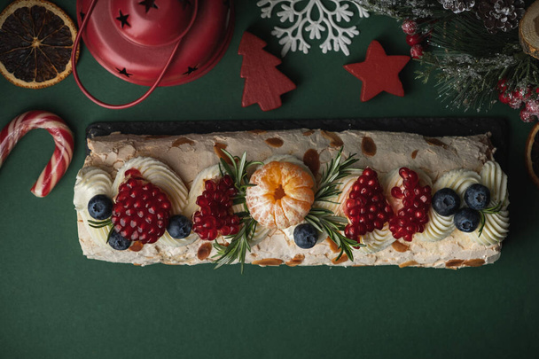 Ρολό μαρέγκας πασπαλισμένο με άχνη ζάχαρη διακοσμημένη με βατόμουρα, ρόδι και νιφάδες αμυγδάλου στο σκούρο πράσινο χριστουγεννιάτικο εορταστικό φόντο. Επίπεδη - Φωτογραφία, εικόνα