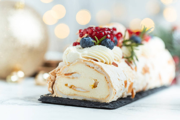 Meringue roll sprinkled with powdered sugar decorated with blueberries, pomegranate and almond flakes on the white background with blurred bokeh lights. Christmas festive pastry - Photo, Image