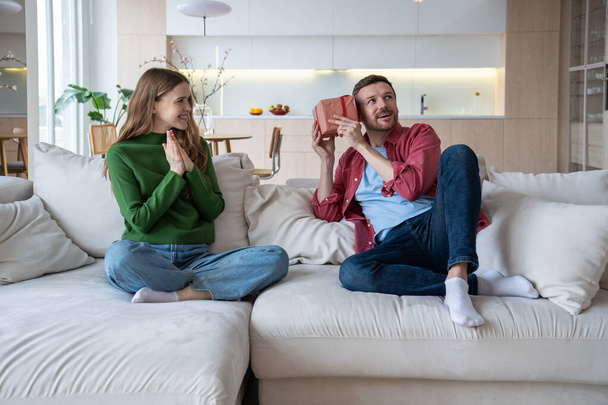 Delighted woman congratulate loving man on holiday, husband shakes present box guessing gift. Happy wife claps hands looking at surprised male. Couple on sofa celebrating birthday, wedding anniversary - Photo, Image