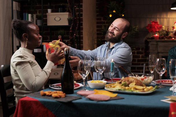 Diverses personnes échangent des cadeaux pour la fête de Noël, répandant la positivité au dîner festif. Joyeux homme donnant boîte cadeau à un ami afro-américain pendant la célébration de décembre. - Photo, image