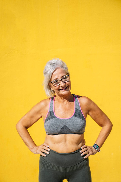 Sportieve oude vrouw in sportkleding poseren en glimlachen in een gele achtergrond - Foto, afbeelding