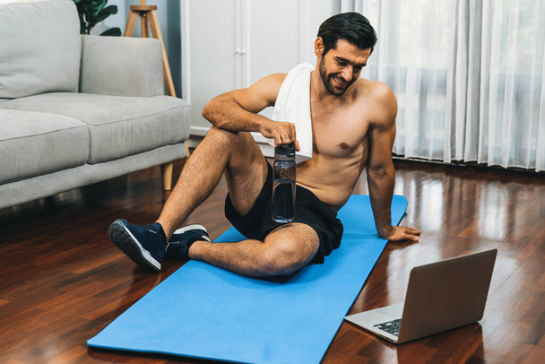 Athletic and sporty man resting on fitness mat during online body workout exercise session for fit physique and healthy sport lifestyle at home. Gaiety home exercise workout training concept. - Photo, Image