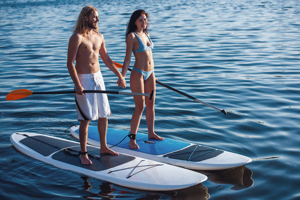 Bella giovane coppia si tiene per mano e sorride mentre SUP surf
 - Foto, immagini
