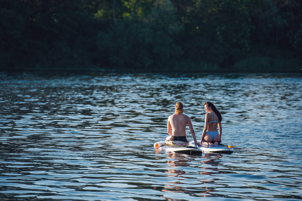 Beau jeune couple parle et sourit tout en étant assis sur des planches SUP sur la rivière
 - Photo, image