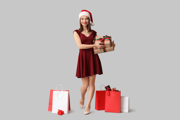Jovem mulher em chapéu de Santa com caixas de presente de Natal e sacos de compras em fundo cinza - Foto, Imagem