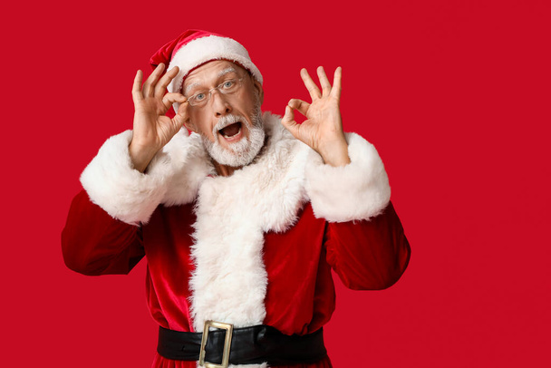 Šokovaný Santa Claus v brýlích ukazující OK gesto na červeném pozadí - Fotografie, Obrázek