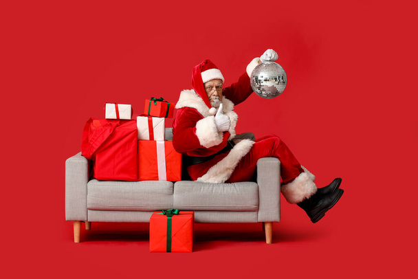 Weihnachtsmann mit Discokugel und Geschenkschachteln mit Daumen-hoch-Geste auf Sofa vor rotem Hintergrund - Foto, Bild