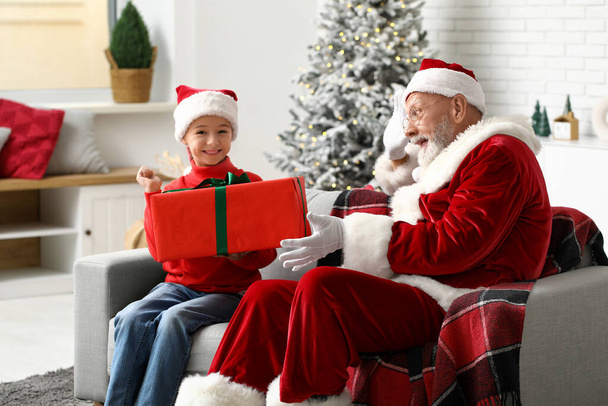 Santa Claus y linda niña con caja de regalo sentado en el sofá en la habitación decorada para Navidad - Foto, imagen