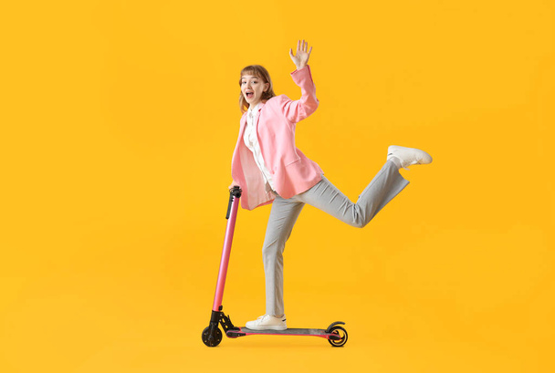 Portrait de femme d'affaires excitée équitation coup de pied scooter et agitant la main sur fond jaune - Photo, image