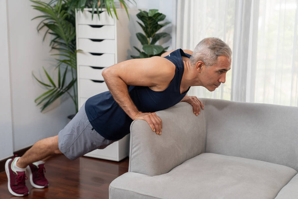 Αθλητικός και δραστήριος ηλικιωμένος άνδρας που χρησιμοποιεί έπιπλα για την αποτελεσματική στόχευση των μυών με push up στο σπίτι άσκηση ως έννοια της υγιούς fit τρόπο ζωής του σώματος μετά τη συνταξιοδότηση. Μύγα - Φωτογραφία, εικόνα