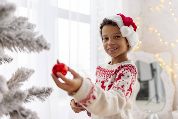 Afro-Amerikaanse tiener jongen in Santa hoed en kerst trui versiert de New Year boom thuis en glimlacht, 10-jarige kind houdt decoratie bal op oudejaarsavond alleen thuis - Foto, afbeelding