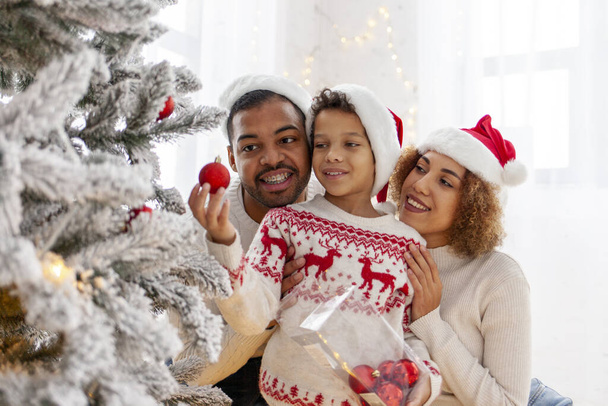 African-American family celebrates Christmas with their child and decorates the Christmas tree at home, 10-year-old boy with his parents in Santa hat holds decorative toy and helps - Photo, Image
