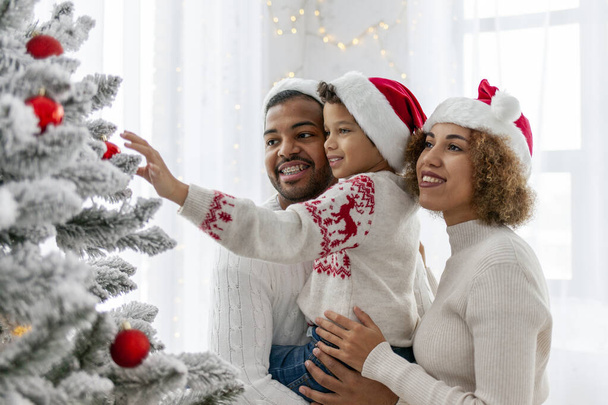 Afroamerikkalainen perhe juhlii joulua lapsensa kanssa ja koristaa joulukuusi kotona, 10-vuotias poika vanhempiensa kanssa Santa hatussa pitää koristeellista lelua ja auttaa. - Valokuva, kuva