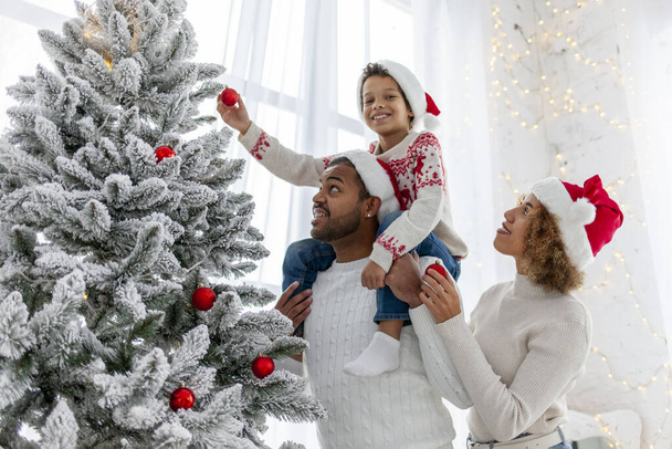 Афро-американская семья празднует Рождество со своим ребенком и украшает елку дома, 10-летний мальчик со своими родителями в Санта шляпе держит декоративную игрушку и помогает - Фото, изображение