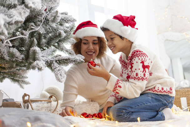 Menino adolescente afro-americano em chapéu de Papai Noel e camisola de Natal decora árvore de Natal com a mãe em casa, a criança mantém o brinquedo de decoração e celebra o Ano Novo com os pais - Foto, Imagem