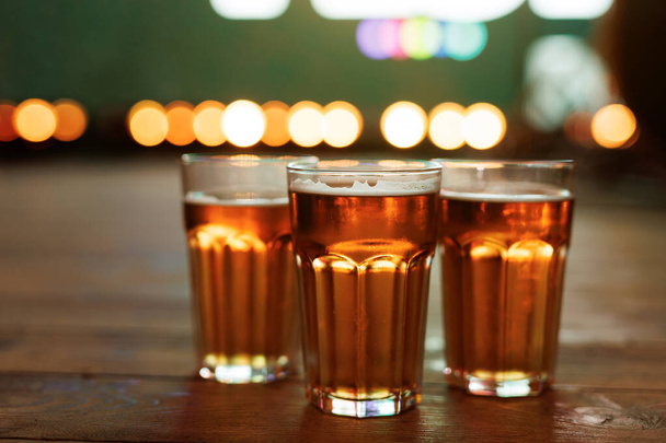 Τρία ποτήρια μπύρας σε ένα ξύλινο μπαρ με πολύχρωμα φώτα bokeh στο παρασκήνιο. - Φωτογραφία, εικόνα