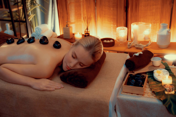 Massage aux pierres chaudes au salon de spa dans un complexe de luxe avec lumière chaude à la bougie, une cliente heureuse profitant d'un massage aux pierres de basalte spa glisse sur le corps avec une chaleur apaisante. Tranquillité - Photo, image