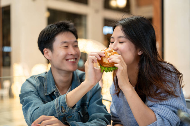 Neşeli ve sevimli genç Asyalı çift, şehirdeki bir restoranda hafta sonu buluşmasında lezzetli bir burger yemekten hoşlanıyor. Buluşma, ilişki, mutlu anlar. İnsanlar ve yemek kavramları - Fotoğraf, Görsel