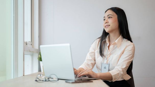 An attractive young Asian businesswoman is looking out the window and daydreaming about her happy career path while working on her laptop computer in the office. - Photo, Image