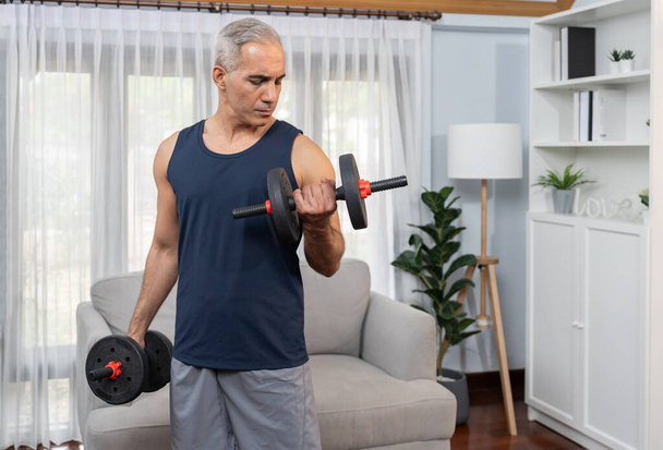 Athletic and sporty senior man engaging in body workout routine with lifting dumbbell at home as concept of healthy fit body with body weight lifestyle after retirement. Clout - Photo, Image