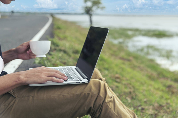 Man hands typing laptop notebook holding coffee cup outside office. Close up man hands using computer laptop sit on green park outdoors lifestyle. freelancer Men using laptop home office technology - Fotoğraf, Görsel