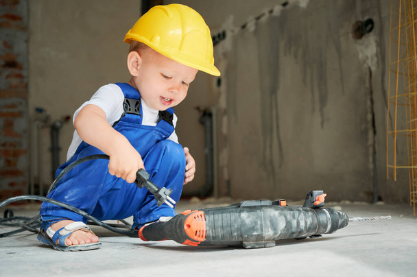 Cute child in safety helmet using rotary hammer drill in apartment under renovation. Kid construction worker playing with demolition drilling machine breaker tool. Home renovation concept. - Photo, Image