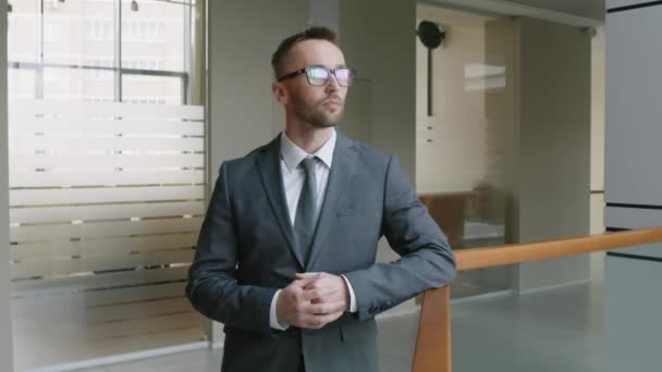 Medium portret van een serieuze blanke zakenman in een bril en grijs pak in een moderne kantoorfoyer en kijkend naar de camera - Video