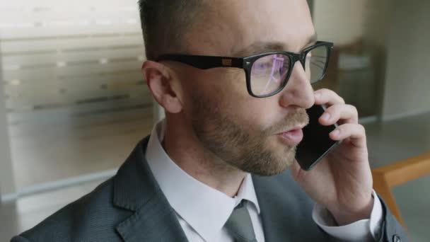 Close-up slowmo shot van blanke mannelijke manager met beugel en bril aan de telefoon met zakenpartners in een moderne kantoorfoyer - Video