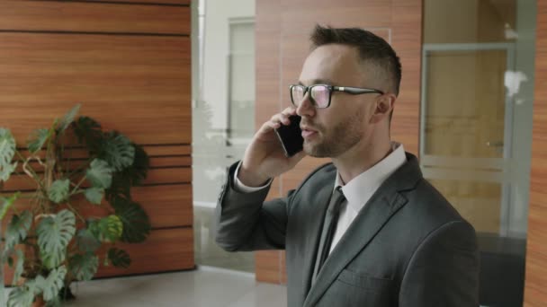 Chest up side shot of Caucasian businessman in glasses and suit speaking on phone in modern office foyer - Imágenes, Vídeo