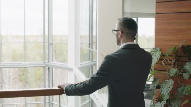 Obloukový střední snímek bělošského obchodníka s brýlemi a šedým oblekem mluvící po telefonu v moderním kancelářském atriu s panoramatickým výhledem - Záběry, video