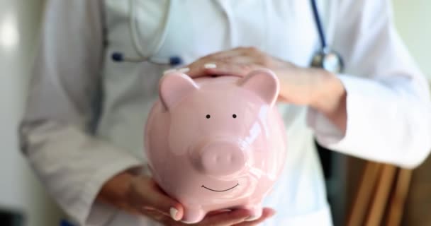 Arzt streichelt rosa Sparschwein mit Münzen Nahaufnahme 4k Film Zeitlupe. Bezahltes medizinisches Versorgungskonzept - Filmmaterial, Video