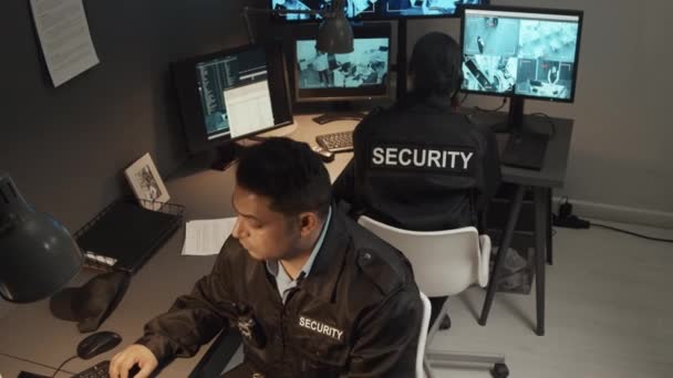 Високий кут зйомки двох різних співробітників безпеки в чорній формі, що працюють і контролюють кілька екранів з відеоспостереженням в кімнаті спостереження - Кадри, відео