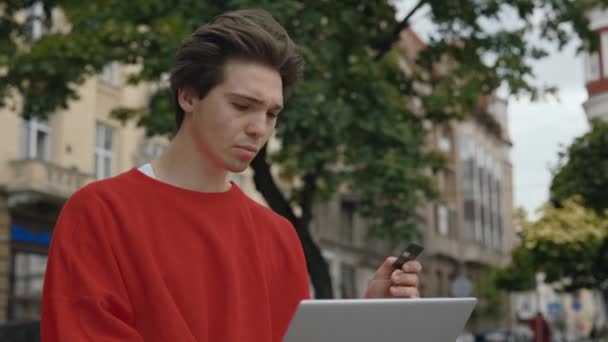 Online Shopping, Payment for Purchases by Credit Card and Laptop. Caucasian Modern Guy Sitting Outside Uses a Laptop and a Banking Card to Pay for Online Purchases or Delivery of Goods - Footage, Video