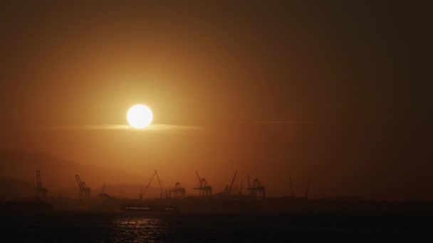 Φορτηγό πλοίο διασχίζει βιομηχανικό λιμάνι το σούρουπο με φόντο το ηλιοβασίλεμα και σιλουέτες γερανού, δημιουργώντας μια γαλήνια σκηνή. - Πλάνα, βίντεο